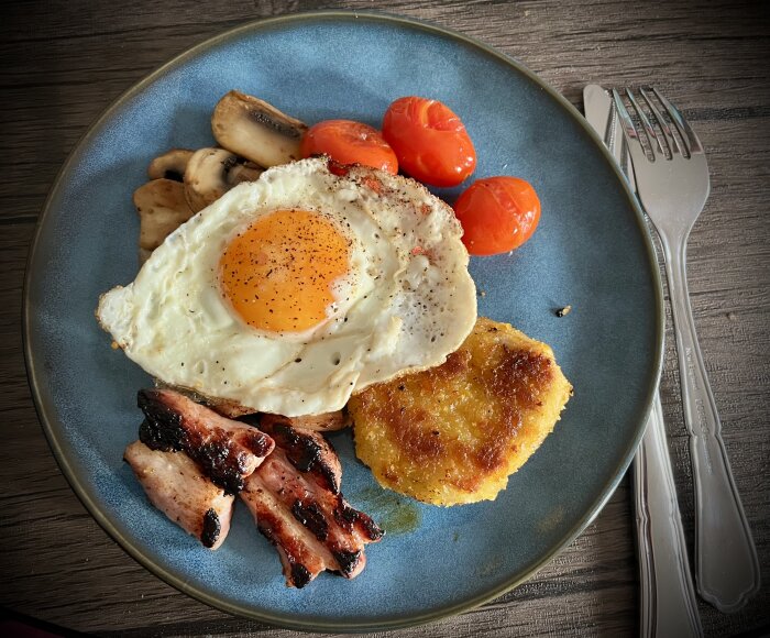 Stekt ägg med krämig gula, stekta champinjoner, röda babytomater, gyllene potatisbulle och krispigt stekfläsk på blå tallrik.