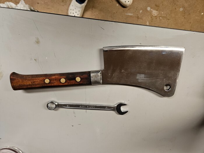 Köttyxa med trähandtag och polerad klinga ligger bredvid en skiftnyckel på en arbetsyta.