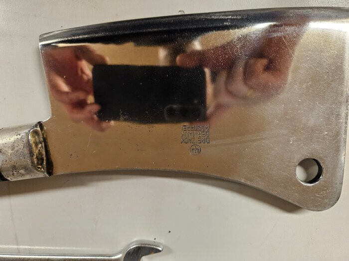Polerad köttyxa med reflektion av en hand och en mobiltelefon, texten "Dexter-Russell, Germany, Stainless" synlig på bladet.