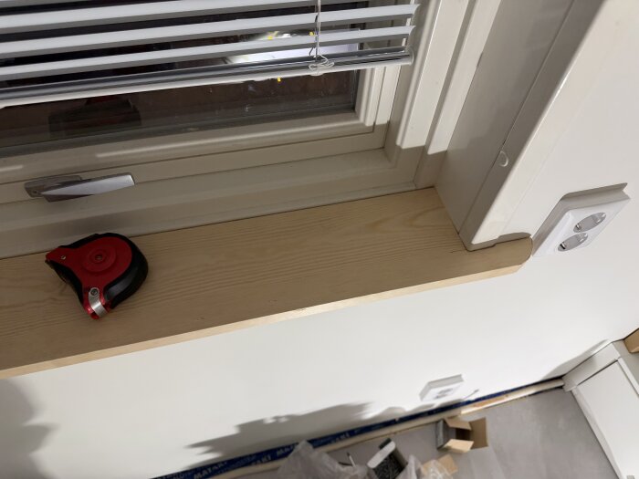 Fönsterbräda med ett måttband och eluttag på en ljus vägg. Fönster med persienner ovanför. Kartonger och verktyg på golvet.