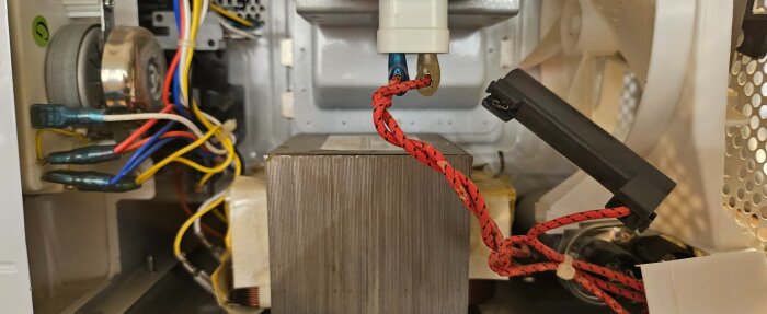 Mikrovågsugn med exponerade elektriska komponenter och kablar, inklusive en svart kontakt med röda sladdar, inuti en öppen hölje.