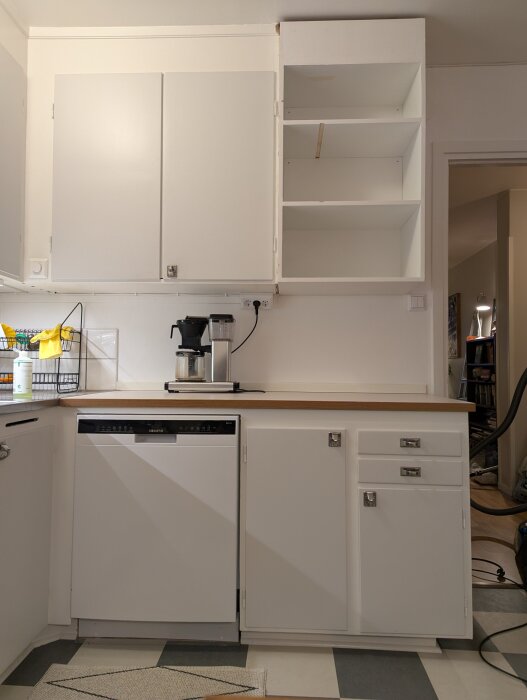 Vitt kök med nytt hemmabyggt öppet överskåp utan dörrar, inrett med flera hyllplan. Under skåpet finns diskmaskin och kaffemaskin.