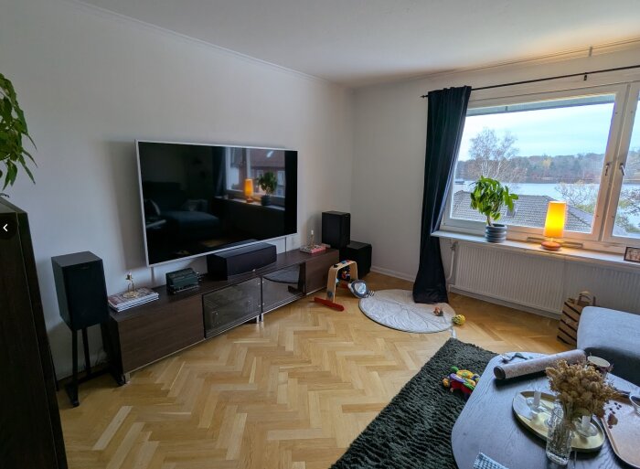 Ett mysigt TV-rum med stor TV, soffbord och fönster med utsikt över ett landskap. Mörka möbler och ljus inredning.