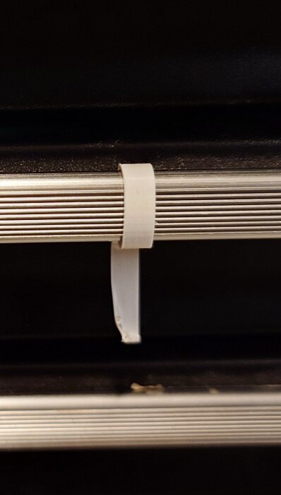 Vit plastklämma på en ribbad metallstång, fäst runt föremålet för att hålla det på plats. Bakgrunden är mörk.
