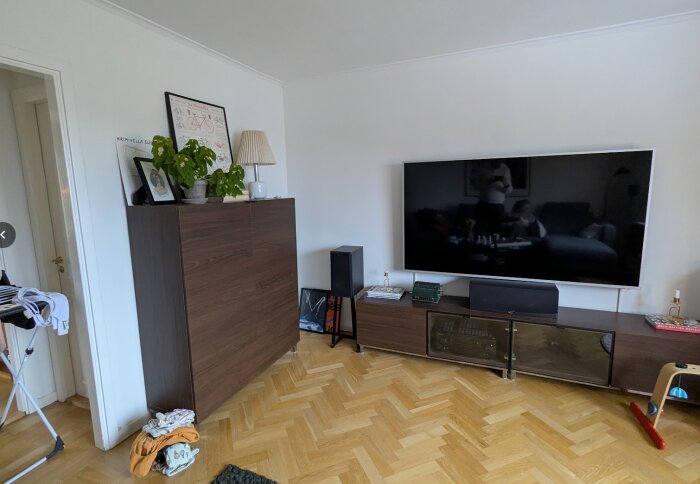 Renoverat TV-rum med stor väggmonterad TV, mörk möbel, växter och lampa ovanpå, ljusa väggar, parkettgolv och en torkställning i hörnet.