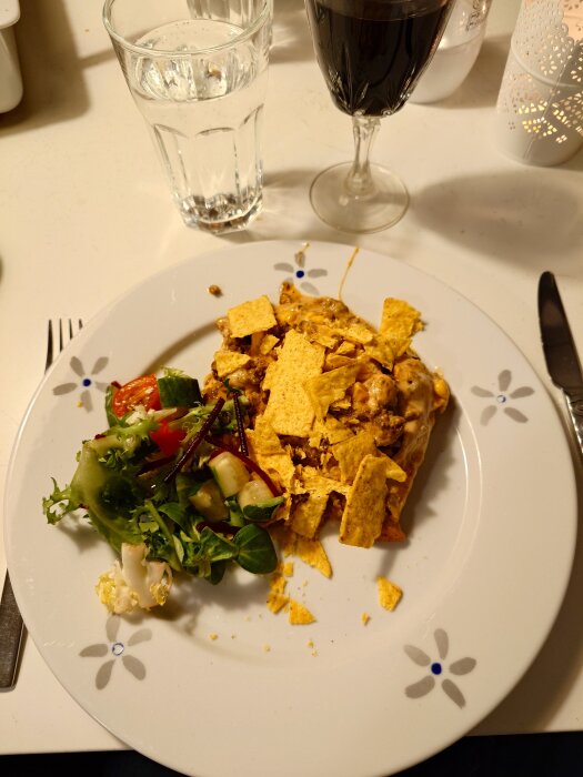 En tallrik med enchiladas toppade med krossade nachos, serverade med grönsallad. Bredvid finns ett glas vatten och ett glas rött vin.