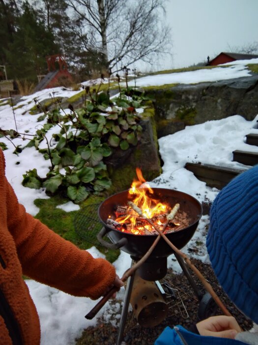 Pinnbröd grillas över en brasa i en trädgård med snö och gröna växter i bakgrunden. Barn med blå mössa syns nära elden.