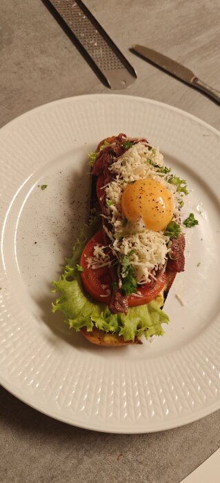 Öppen smörgås med sallad, tomatskivor, kött, strimlad ost och rå äggula på en vit tallrik på ett bord.