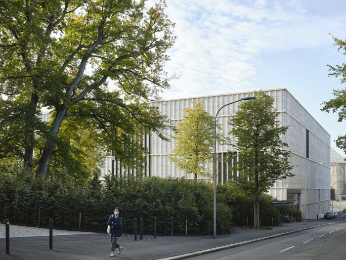 En modernistisk byggnad omgiven av träd och växter, med en person som går på gatan framför, under en klar himmel.