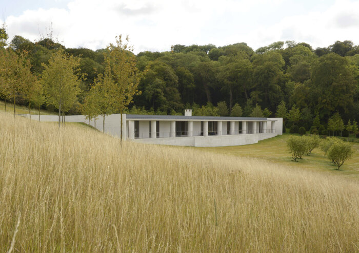 Modernt hus i betong med pelare, omgiven av gräs och skog. Långa raka linjer och minimalistisk design.