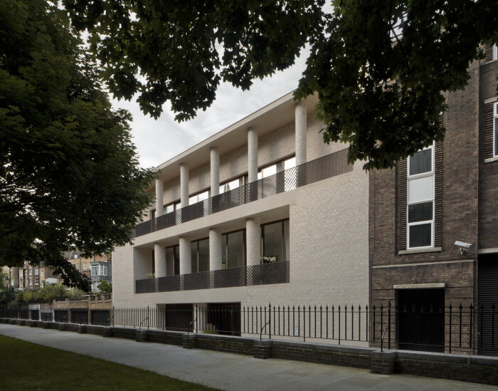 Modern byggnad med två våningar, tegel och pelare, omgiven av träd. Staket och trottoar i förgrunden. Minimalistisk arkitektur.