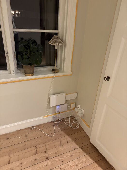 Hörna med teknikutrustning och trassliga kablar under ett fönster med krukväxt och lampa på fönsterbrädan.