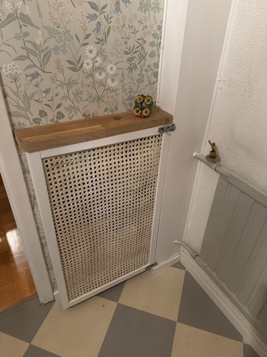Flätat träradiatorskydd med trälist upptill, mot mönstrad tapet och schackrutigt golv.