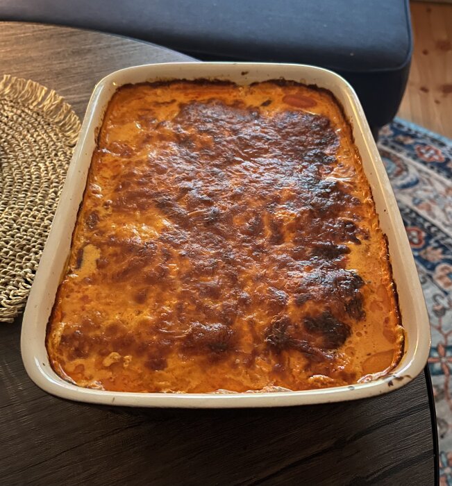 En gratängform med nygräddad lasagne på ett träbord, bredvid en flätad bordstablett.