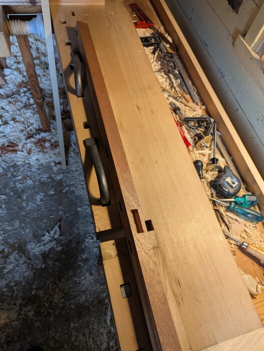 Arbetsbänk med trädelar av sängram, verktyg och träspån. Skåror och beslag synliga.