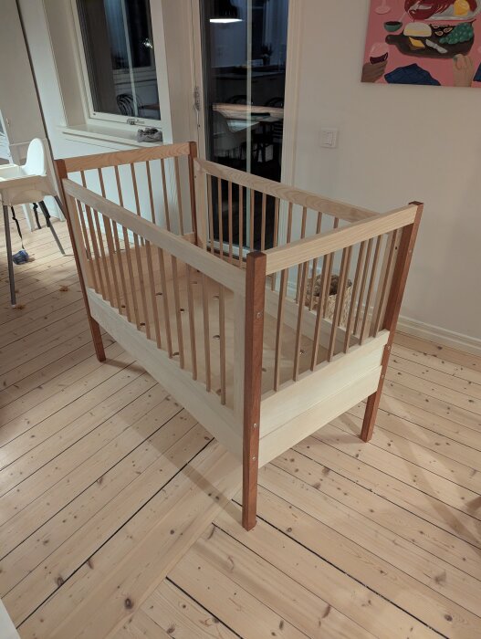 En träspjälsäng under konstruktion i ett ljust rum med trägolv och ett färgglatt konstverk på väggen.