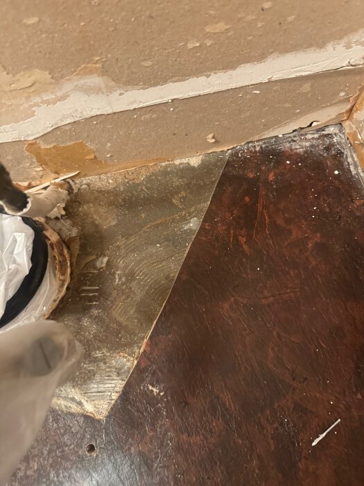 Plastmatta delvis upplyft från mörkt golv, visar slät undersida och gammal betongvägg, del av renovering av gästtoalett i hus från 1978.