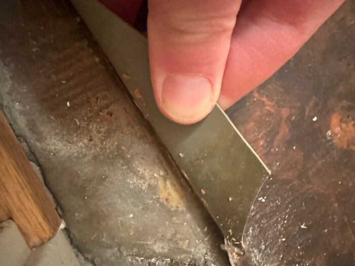 En hand håller upp en bit av en grön plastmatta som är fastlimmad på ett betonggolv i en gästtoalett under renovering.