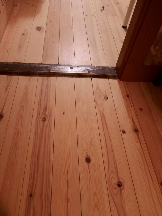 Furugolv med två olika ytbehandlingar på var sida om en tröskel, med synliga träknotor och ljus nyansskillnad.