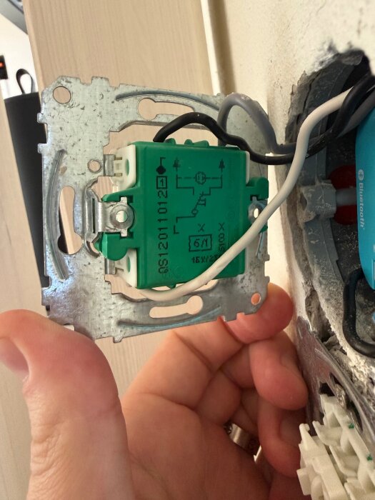 En handen håller en grön elektrisk komponent med kopplingar, monterad på en metallplatta, i en pågående installation i en vägg.