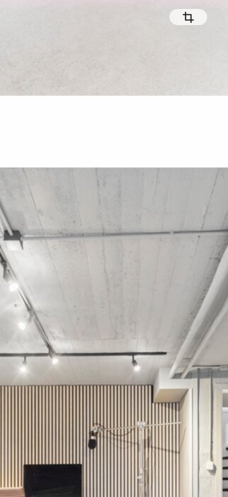 Innertak med brädliknande struktur och synliga rör på undersidan, i ett rum med en detaljrik trävvägg och infällda spotlights.