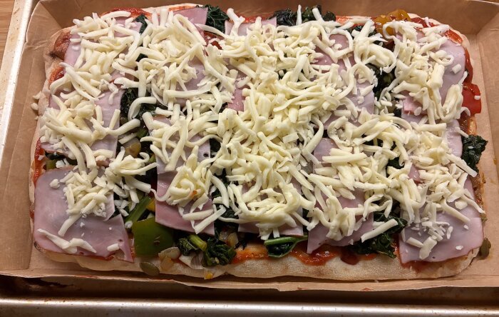 Hemlagad pizza med tomatsås, pålägg av grönsaker, skivad skinka och toppad med riven ost på ett bröd.