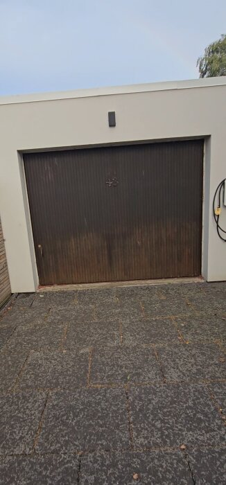Sliten vipport i trä på garage med vit putsad fasad, omgiven av stenbelagd uppfart.