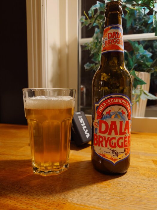 Flaska och glas med öl från Dala Bryggeri på ett bord, gröna växter i bakgrunden.