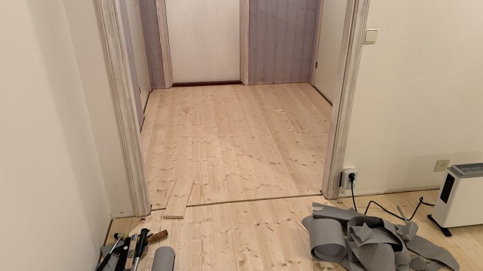 Massivt grangolv under installation med mellanrum för tröskel; verktyg och material synliga, kabelansluten värmare i hörnet.
