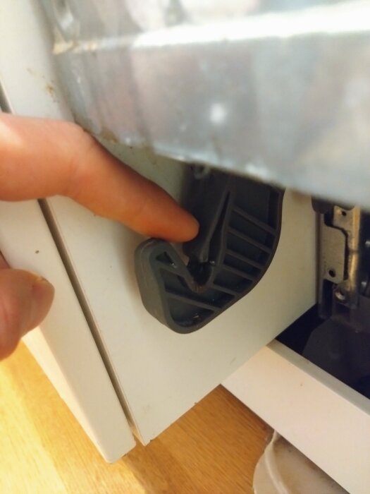 Finger trycker in stift på plastkrok bakom diskmaskinsfront, nära dörrens nedre del, för att lossa fronten.