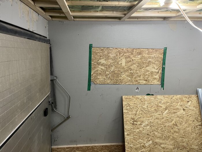 Garagevägg med monterade träskivor och en metallarm kopplad till garageporten i en renoveringsmiljö.
