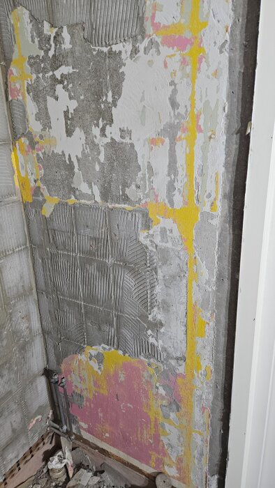 Vägg med avslitet gips, synlig gammal lim och målarfärgslager i rosa och gult, rör i hörn och byggmaterial på golv.
