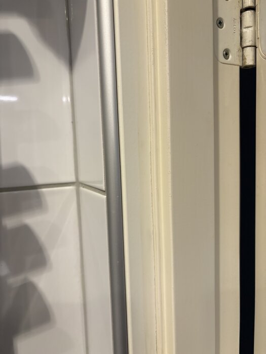 Fuktskadad golvlist mot dörrkarmen i badrummet, intill kakelväggen och dörrgångjärn. List verkar limmad.