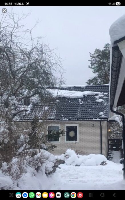 Snö täcker taket på ett hus med svart takpannor, omgiven av träd och buskar, föreslår behovet av snörasskydd.