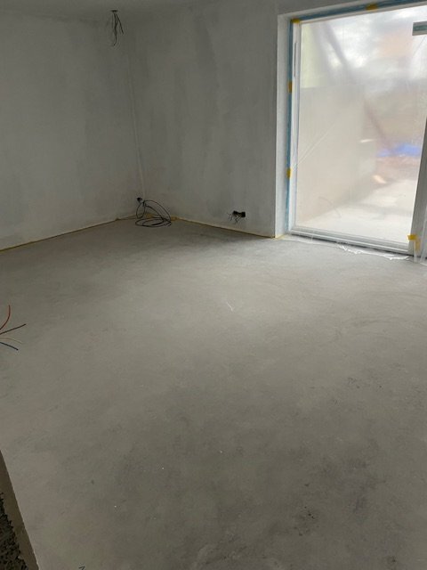 Obehandlat betonggolv i ett rum under konstruktion med stora fönster som släpper in dagsljus, kablar löper längs väggarna.