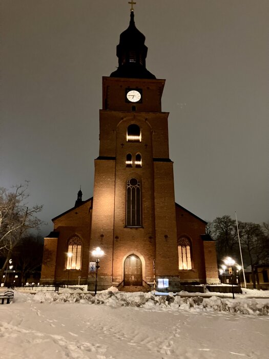 Tegelbyggnad av en kyrka i kvällsbelysning, omgiven av snö. Klocka synlig på kyrkans torn.