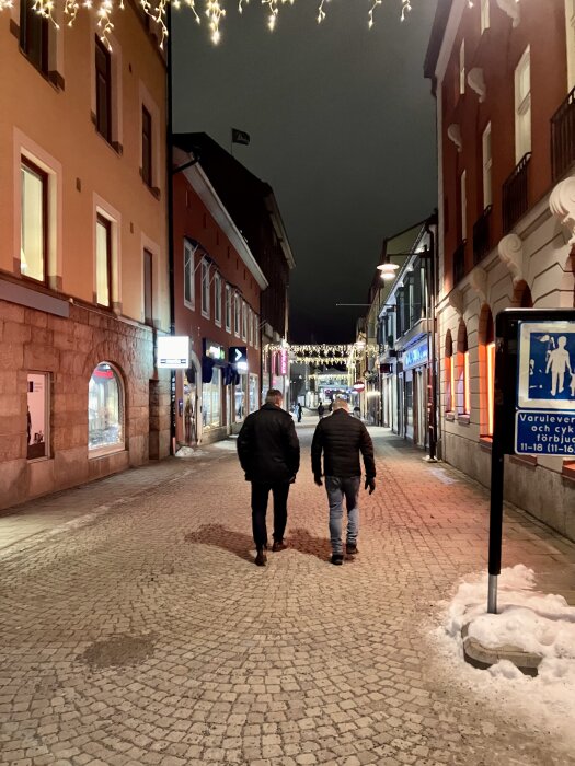 Två personer går på en upplyst stenlagd gata kantad av butiker och dekorerad med ljusslingor, en vinternatt.