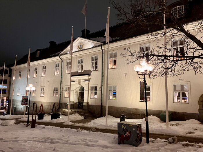 Upplyst byggnad i snöigt landskap med flaggstänger och gatulampor på kvällen, träd utan löv i förgrunden.