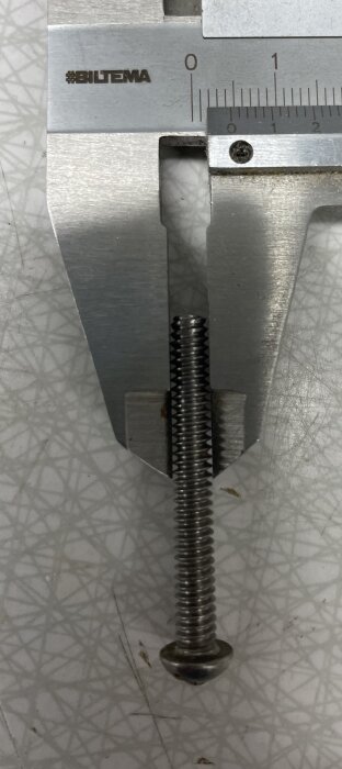 Metallskruv mätt med skjutmått på ett rutigt underlag.
