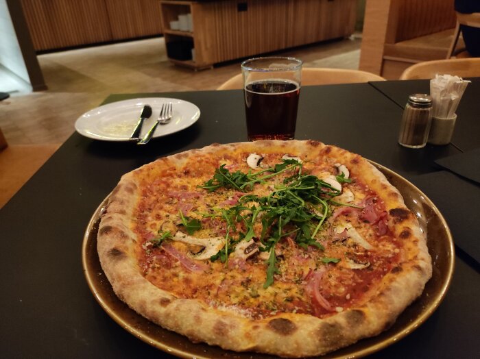 Pizza med ruccola och olika toppings på bord i restaurangmiljö. En läsk står bredvid tillsammans med bestick och servett.