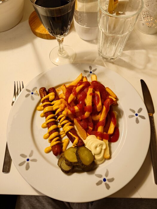 Tallrik med pommes frites och korv toppad med ketchup och senap, serverad med gurka och en klick majonnäs, glas vin i bakgrunden.