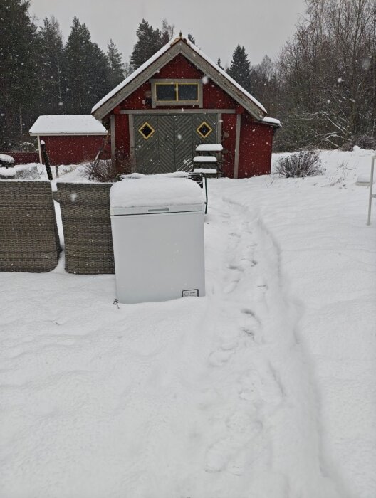 Snöbelagd gård med rött uthus, staket, och en kall frysbox omgiven av sittmöbler. Fotspår leder bort från kamera, skog i bakgrunden.