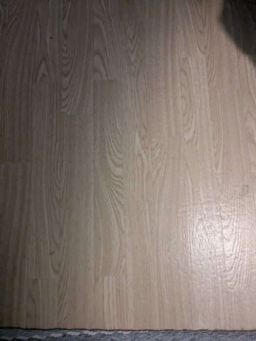 Bild av vinylmatta på spånskiva, mönstrad som trä, del av golvrenovering. Frågor om ålder och säkerhet, ingen synlig svart lim.