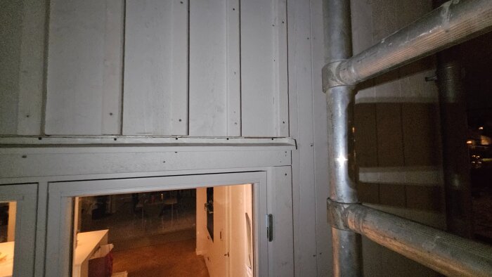 Fasad av ett hus med träpanel och fönster, belyst på kvällen. Byggnadsställning synlig till höger.