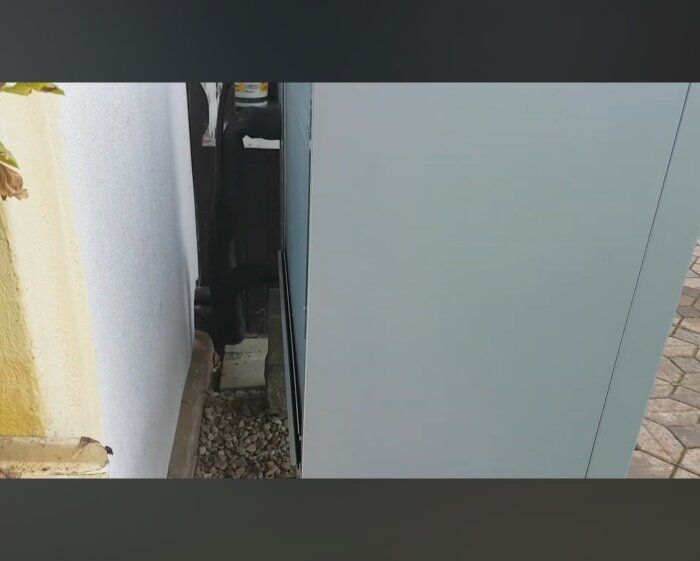 Utrymme mellan fasadvägg och värmepump, med exponerade rör mot grusbotten.