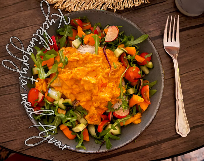 Tallrik med kycklinggryta täckt med ajvar, omgiven av färgglad sallad med gurka, tomat och rucola. Gaffel placerad till höger om tallriken.