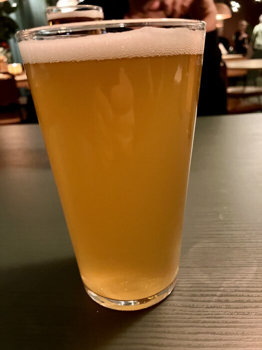 Ett glas med gyllene öl och skum på toppen står på ett mörkt bord, med en suddig bakgrund av restaurangmiljö.