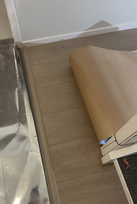 Laminatgolv med en smal ändbit på tre cm mot en vit vägg, med pappersark delvis utrullad över golvet.
