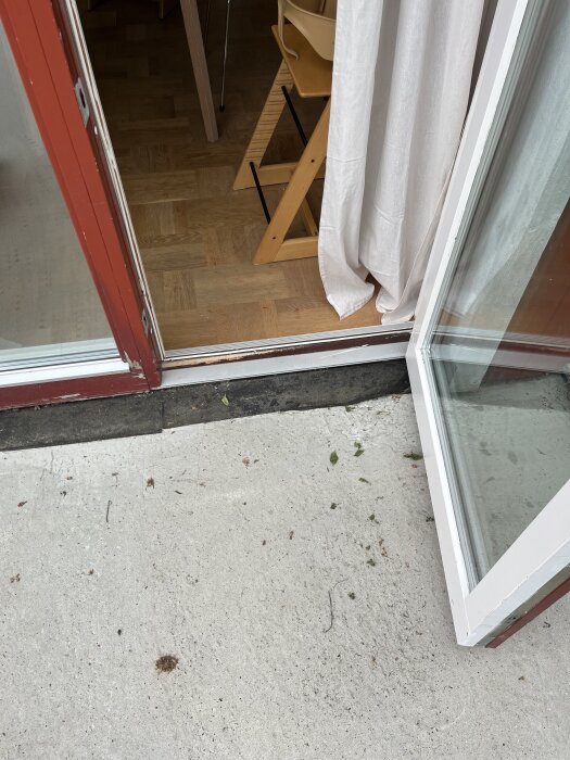 Öppen dörr mot ett rum med parkettgolv, vita gardiner och en trätrappstol. Betonggolv syns utanför dörren.