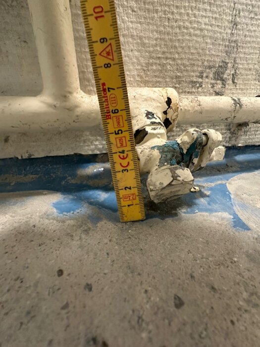 Gammalt elementrör med synlig termostat och måttband mot blå tätning och betonggolv.